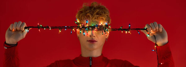 Стильный молодой человек стоит на ярком красном фоне, игриво держа нить красочных рождественских огней.