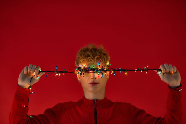 Стильный мужчина, одетый в праздничный наряд, игриво держит верёвку ярких рождественских огней на лице..
