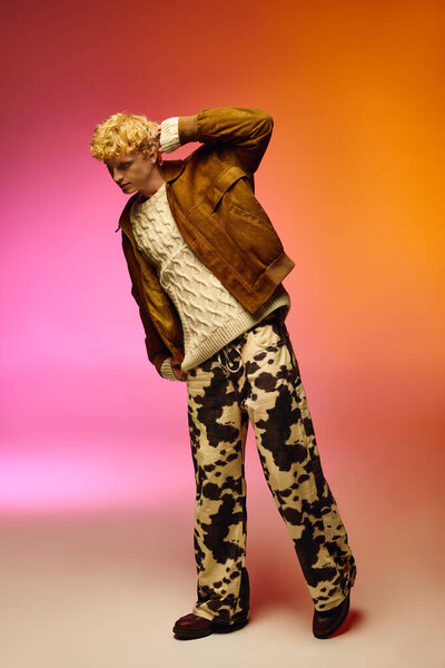 A stylish man showcases holiday fashion trends with a cozy knit sweater and a brown leather jacket. He pairs it with unique patterned pants, creating a chic festive look against a vibrant backdrop.