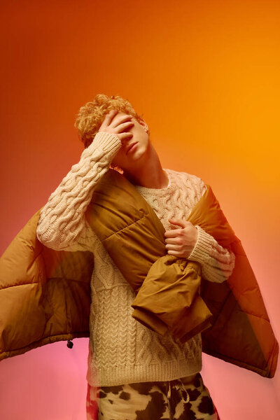 Молодой человек демонстрирует модную праздничную моду, надевает уютный свитер и держит стильный пиджак, окруженный яркими праздничными красками. Настройка излучает тепло и праздничный дух на Рождество.