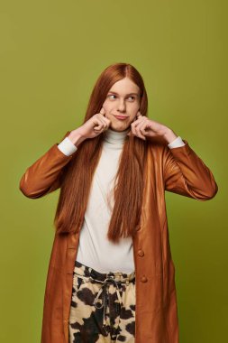 Uzun kızıl saçlı genç bir adam şık bir kıyafet giyerken şakacı bir şekilde yanaklarını çekiyor..