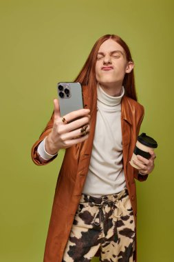 Uzun kızıl saçlı genç bir adam elinde kahve fincanı, elinde bir selfie tutarken neşeli bir poz veriyor..