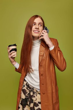 Uzun kızıl saçlı genç bir adam elinde kahve ve telefonda sohbet ederken gülümsüyor..