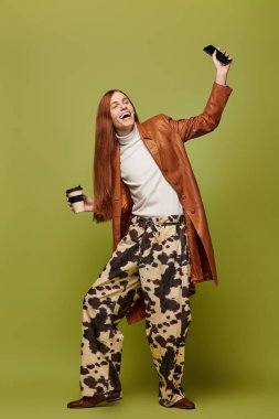 Young man with long red hair wears trendy outfit while holding coffee and smartphone, smiling. clipart