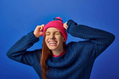 Happy young man with long red hair laughs in a colorful hat against a blue background. clipart