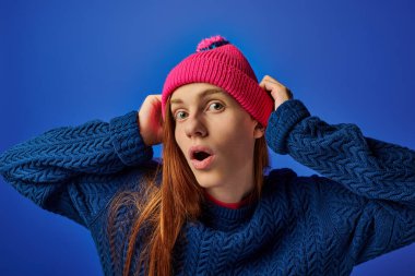 A young man with long red hair, surprised, wears a pink beanie against a blue backdrop. clipart