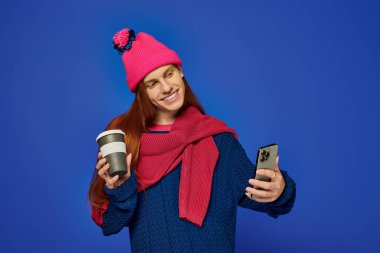 Young man with long red hair smiles while holding coffee and checking his phone in winter gear. clipart