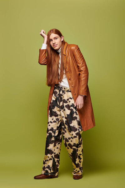 A young man with long red hair poses in unique attire against a green background.