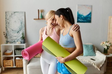 İki genç kadın evde yoga antrenmanına hazırlanırken kahkaha ve şefkat paylaşıyor..