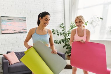 İki genç kadın yoga minderlerini yeşilliklerle dolu, modern, havadar bir yerde mutlu bir şekilde hazırlıyorlar..
