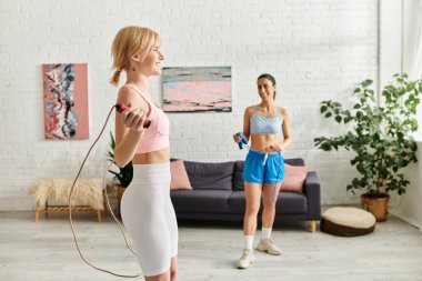 Two young women enjoy a playful workout, laughing together in their cozy living space. clipart