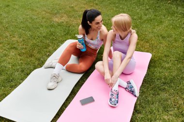 İki genç kadın yoga minderlerinde rahatlar, güneşli bir parkta birbirlerine eşlik ederler..