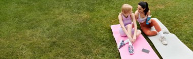 Two young women share smiles and laughter while relaxing on yoga mats in a green park. clipart