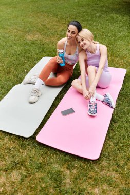 İki kadın yeşil bir parkta yoga minderlerinde gülüyor, neşe ve dostluk gösteriyorlar..