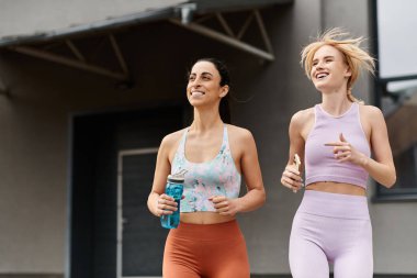 İki genç kadın spor kıyafetleriyle neşeli bir şekilde koşuyor, egzersiz sırasında gülüşlerini ve pozitifliklerini paylaşıyorlar..