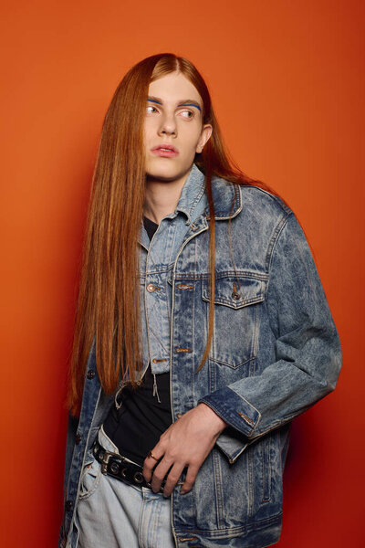 Молодой человек стоит с длинными рыжими волосами, демонстрируя различные эмоции перед оранжевой стеной.