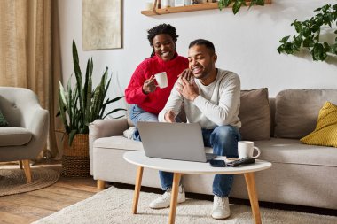 A loving couple enjoys quality time together, laughing and sipping coffee as they relax at home near laptop clipart