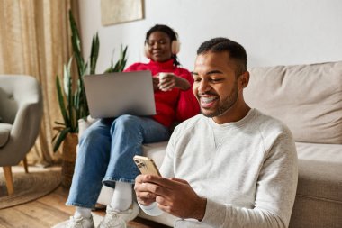 A joyful African American couple relaxes indoors in warm sweaters, using gadgets clipart