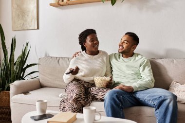 A loving couple enjoys each others company, sharing smiles and popcorn in their cozy living room. clipart