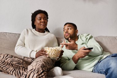 A happy couple enjoys popcorn together while relaxing on a cozy couch at home, embracing winter. clipart