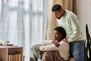 African American man comforting his worried girlfriend in their cozy living room clipart