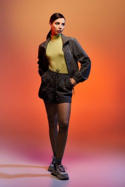 Tarz sahibi genç bir kadın sıcak bir arka plana karşı şık kıyafetler içinde poz veriyor..