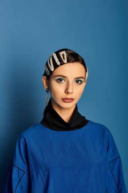 Genç bir kadın şık kıyafetlerini ve aksesuarlarını canlı, mavi bir stüdyo ortamında sergiliyor..