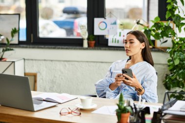 Kumral saçlı genç bir kadın bitkilerle dolu canlı bir çalışma alanının ortasında telefonuna odaklanmış..