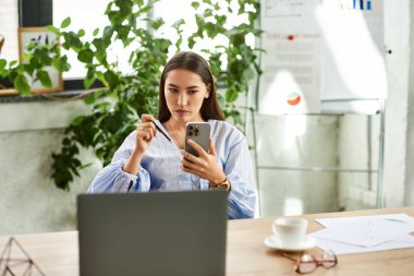 Kumral saçlı genç bir kadın iş yerinde yeşilliklerle çevriliyken telefonuna odaklanıyor..