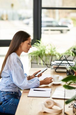 A young woman with long brunette hair sits at a wooden table, sipping coffee and using her phone. clipart