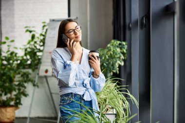 Brunette woman stands by large windows, sipping coffee and chatting happily on the phone. clipart