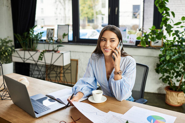 Young brunette woman smiles while talking on the phone in a stylish office setting.