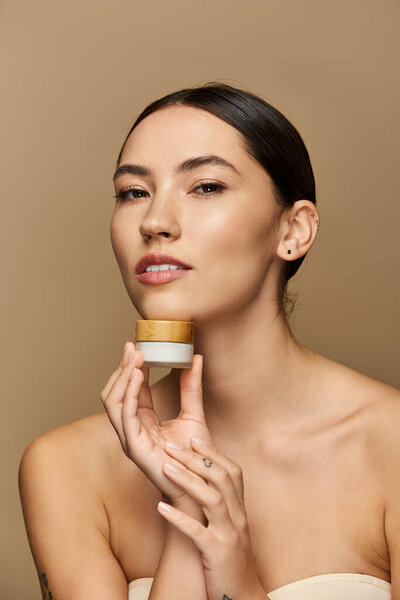 Молодая женщина держит продукт по уходу за кожей, подчеркивая ее сияющий цвет лица в спокойных тонах.