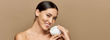 Genç esmer kadın, minimalist bir arkaplanda cilt bakımını neşe ve güvenle sergiliyor.