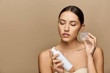 Genç bir esmer, elinde bir ürün ve konteynır tutarken cilt bakımı yapıyor..