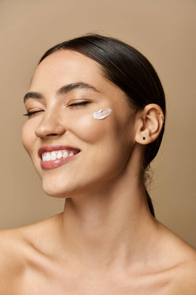 Beautiful brunette woman applies cream on her cheek, radiating confidence and joy while smiling.