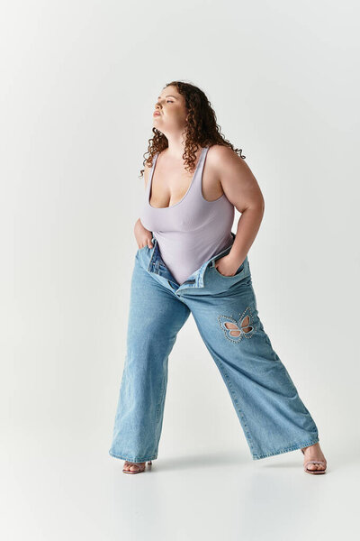 Young woman confidently poses in oversized jeans and a fitted top against a minimalist backdrop.
