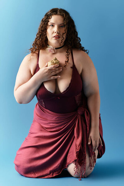 A chic young woman kneels gracefully against a blue background, radiating confidence.