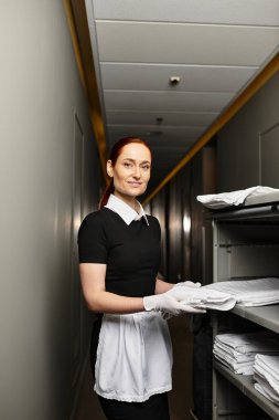 A beautiful woman in a uniform carefully arranges folded linens in a hotel corridor. clipart