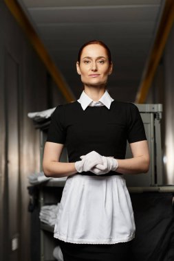 A young woman in uniform stands proudly in a sleek hotel corridor, showcasing her professionalism. clipart