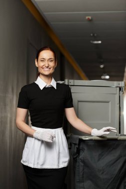 Temiz bir koridorda çöp kutusunun yanında duran üniformalı genç bir kadına parlak bir gülümseme..