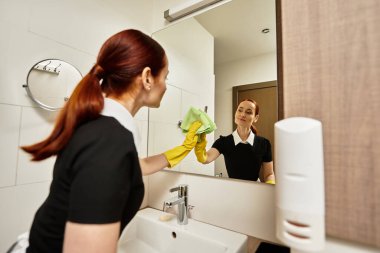 Genç bir kadın, banyo aynasını parlak bir alanda yeşil bir bezle temizlerken gülümsüyor..
