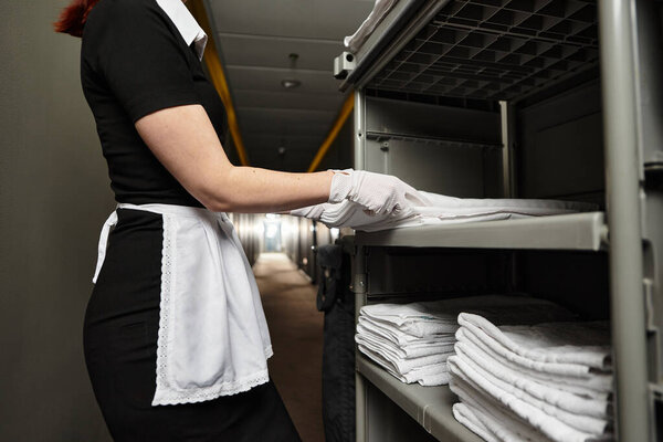 A beautiful young woman in uniform organizes plush towels on shelves, enhancing hotel aesthetics.
