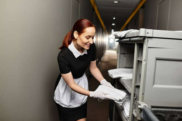 A young woman in a uniform smiles while organizing fresh linens on a cart in a hotel hallway.