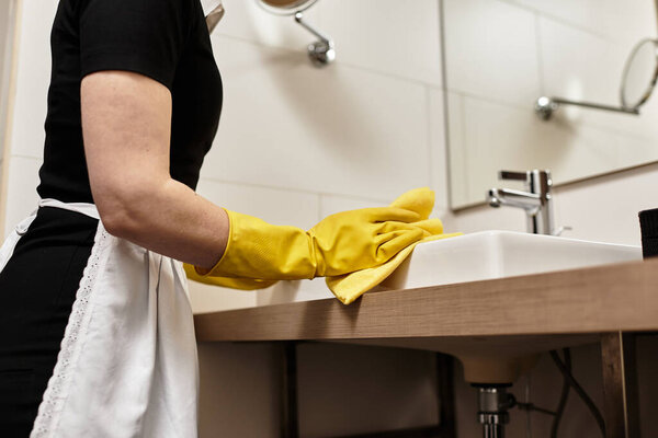 Светлая ванная комната, где молодая женщина тщательно чистит раковину желтыми резиновыми перчатками.