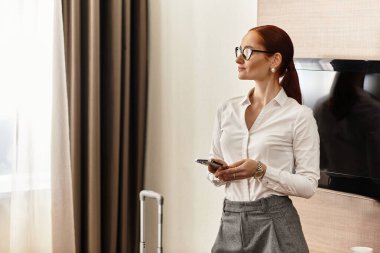 Confident young woman dressed in stylish attire stands in a hotel room, glancing at her phone. clipart