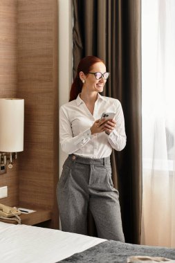 Güzel bir kadın pencerenin kenarında duruyor, rahat bir otel odasında telefonunu kontrol ediyor..