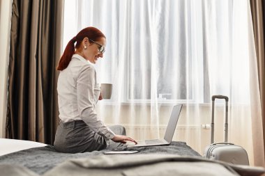 Genç bir kadın, sıcak bir otel odasında yatağında kahvesini yudumluyor ve laptopunu kullanıyor..