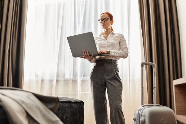 Beautiful young woman focuses on her laptop in a stylish hotel room, preparing for a trip.