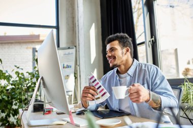 Modern ofis ortamında kahvesini yudumlarken tasarım işleriyle uğraşan çekici bir adam.